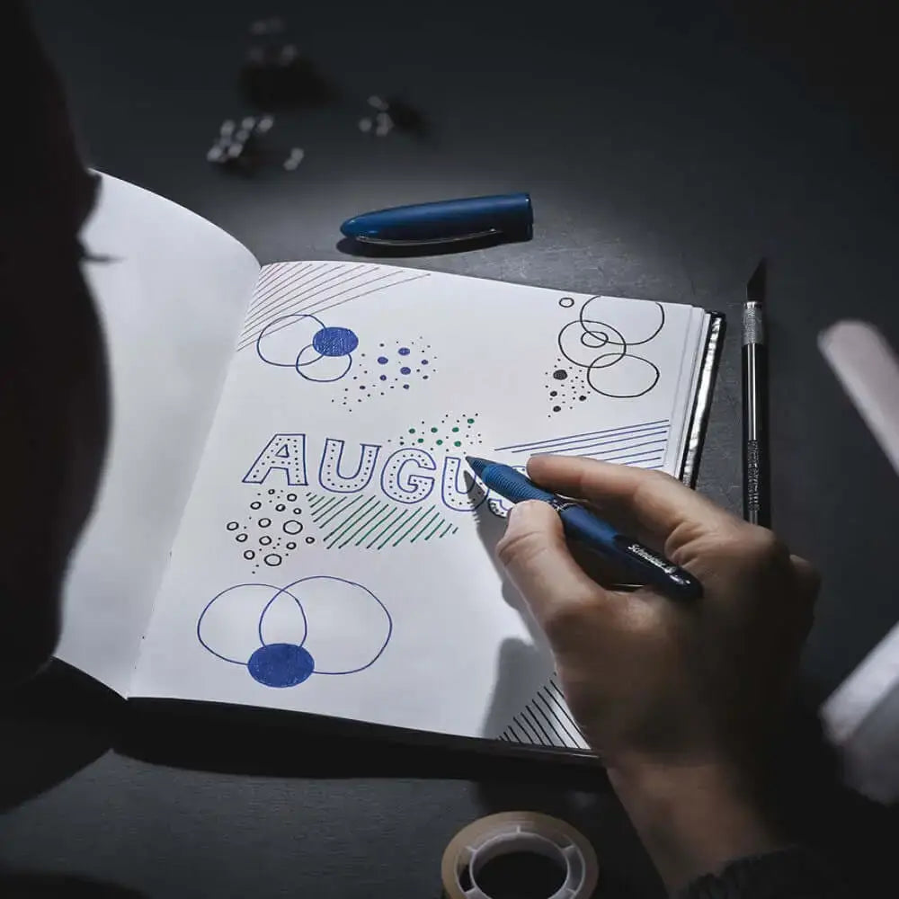 Person using Schneider One Business 0.6MM Roller Ball Pen to write in a notebook showcasing designs for August.