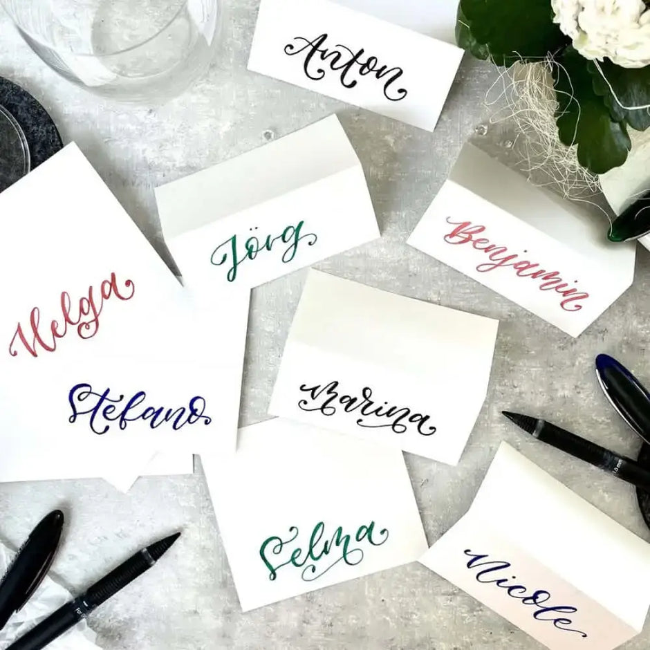 Handwritten names on decorative cards beside pens, showcasing elegant calligraphy styles.