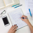 Person writing in a notebook using a Schneider Slider Rave XB Ball Point Pen with glasses and a mobile phone nearby.
